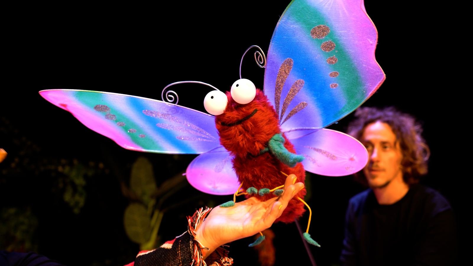 A vivid pink and blue butterfly puppet perched on a performer's hand.