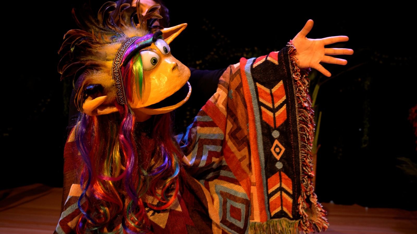 A brightly costumed Gumba puppet with a rainbow-feathered headdress, mid-performance on stage.