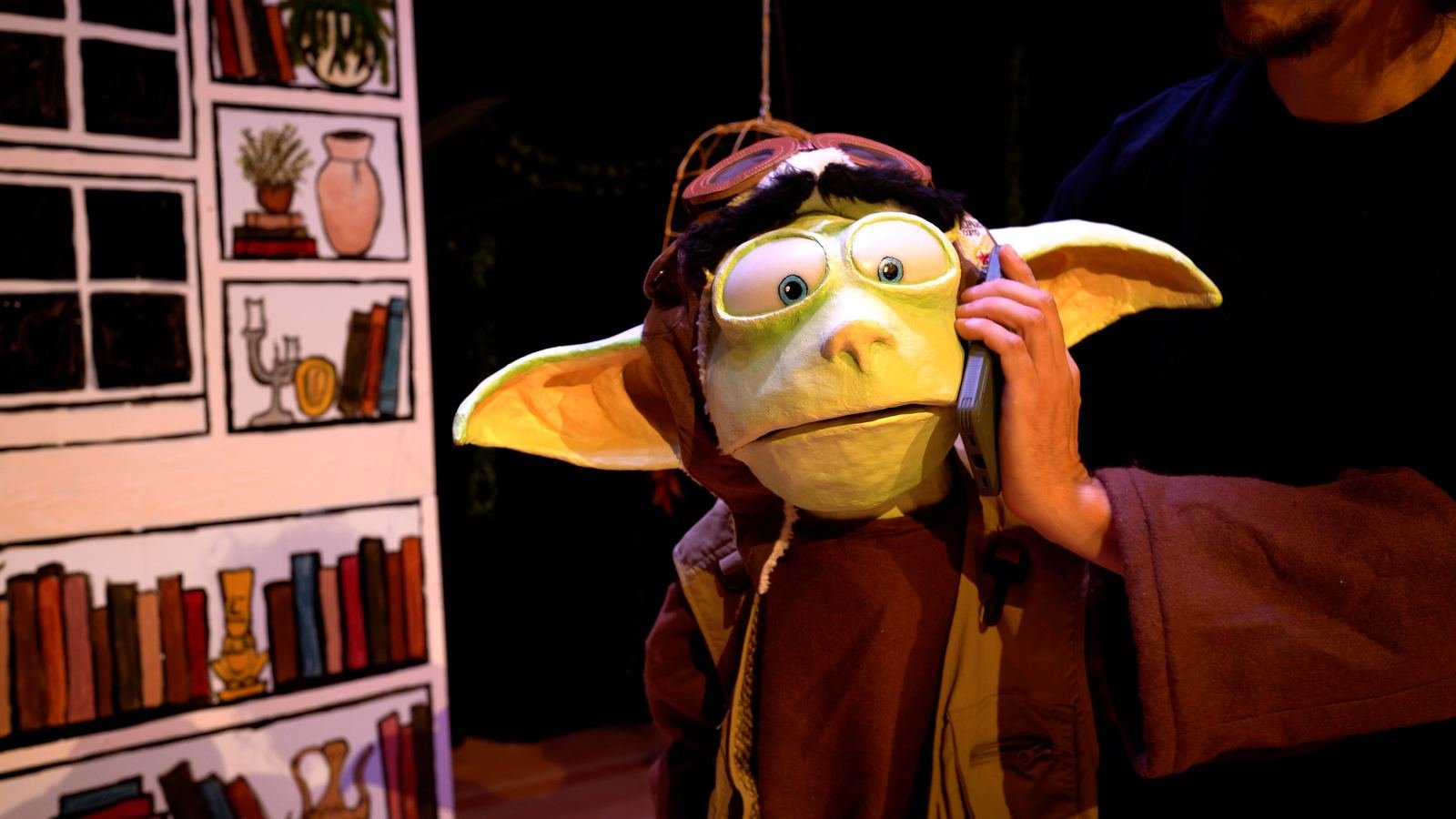 A goggled Gumba puppet holding an old telephone to its ear, mid-scene.