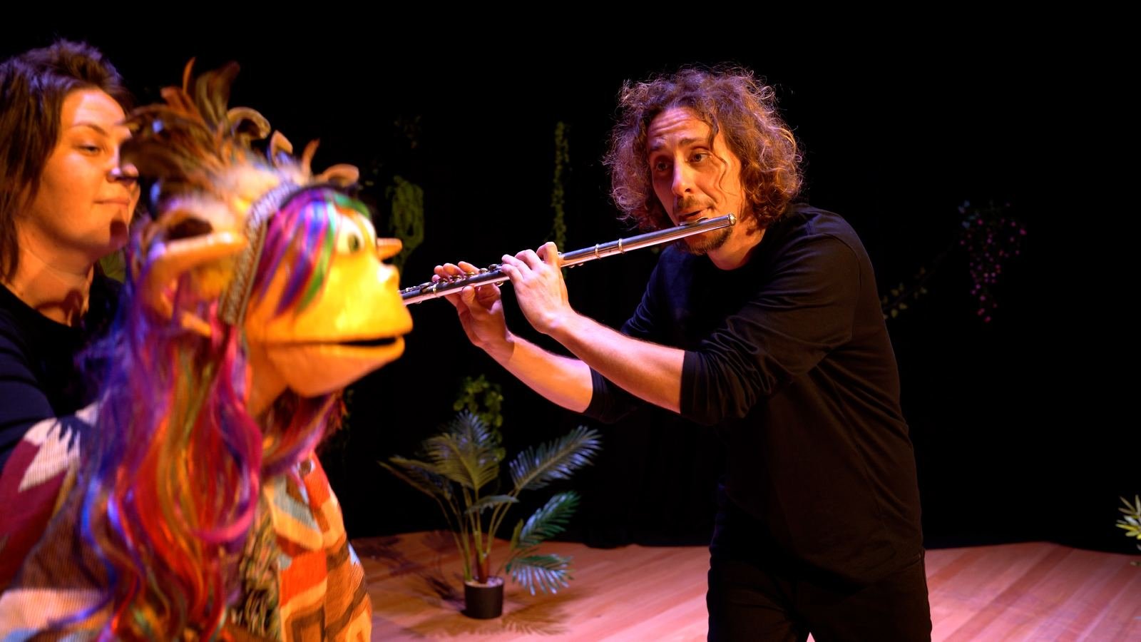 A performer plays flute beside a colourful long-haired puppet on stage.